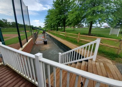 Wooden ramps leading to a baseball field, with a fence and trees in the background on a sunny day.