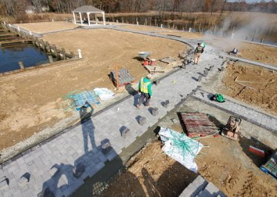 Construction workers building a paved walkway near a body of water with a gazebo in the background.