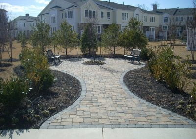 Paved garden path lined with plants, leading to benches and trees, surrounded by suburban houses.