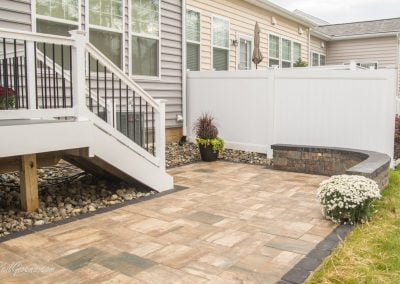 A backyard with stone tiles, white railing stairs, potted plants, and a white fence along a house with beige siding.