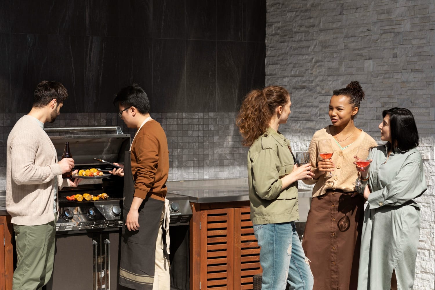 2148965322 Five people enjoying an outdoor kitchen barbecue; two grilling food while three chat and hold drinks, all soaking up the lively atmosphere.