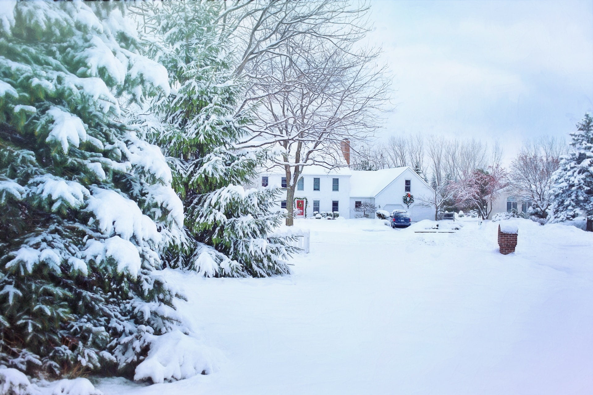 A snow-covered landscape with a house, large evergreen tree, bare trees, and a car parked in the driveway.