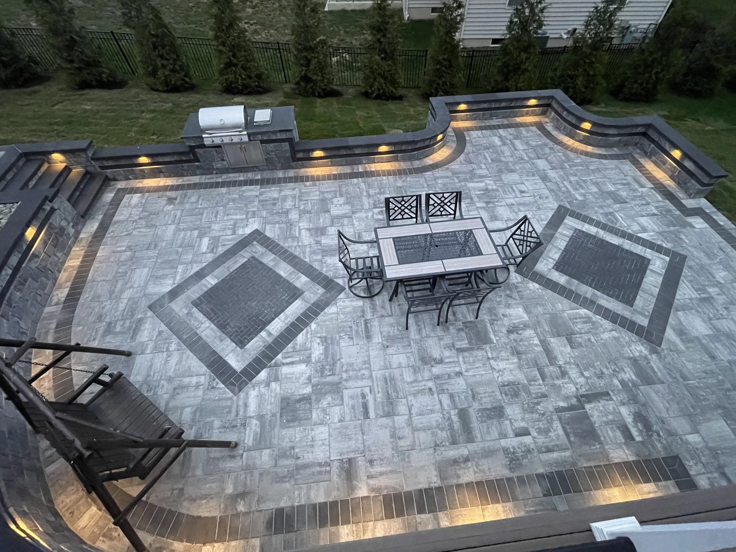 IMG_4044-scaled A well-lit outdoor patio with a dining set, built-in grill, and geometric-patterned stone flooring showcases exquisite hardscape design, bordered by trees.