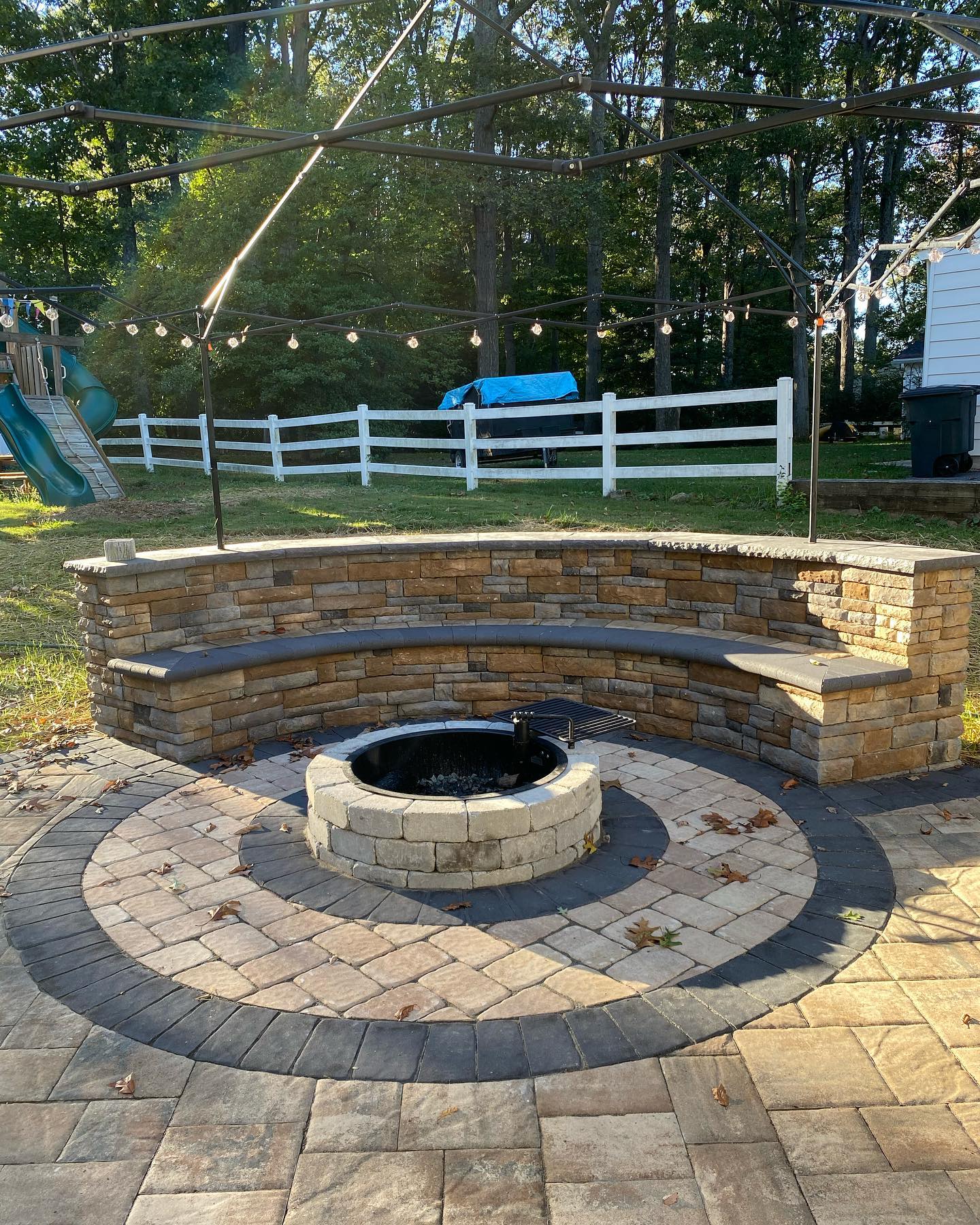 Outdoor stone fire pit with curved seating and string lights in a backyard featuring a white fence, greenery, and an inviting outdoor fireplace.