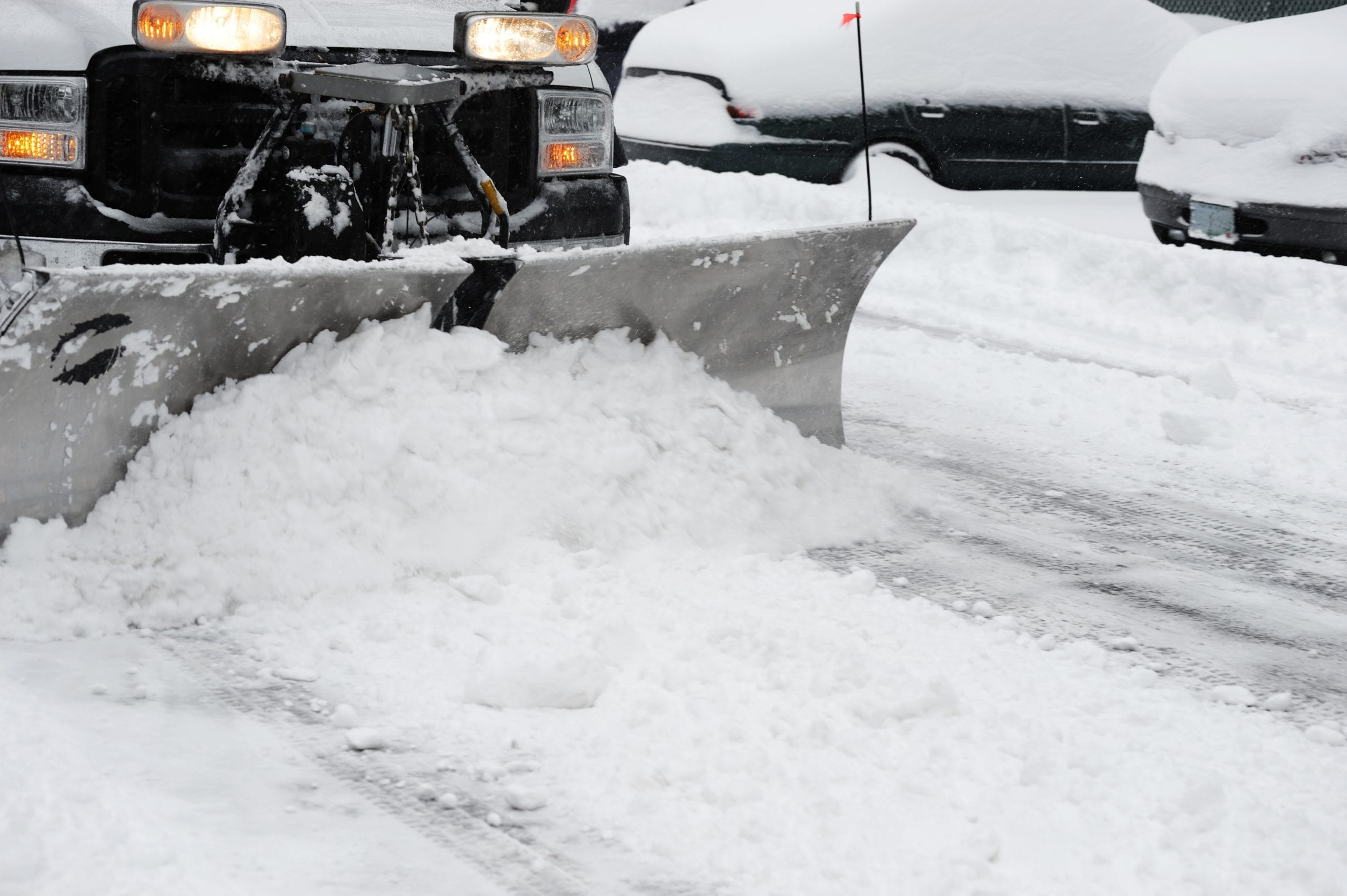 snowplow removing snow on the street after blizzard snow removal companies Snowplow clearing a snow-covered street, with parked cars partially buried in snow in the background.