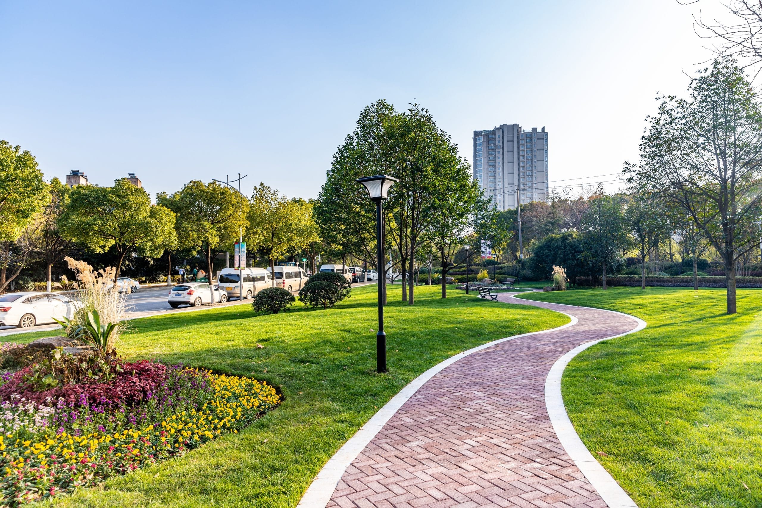 A curved brick path in a sunny park with trees, flowers, and a tall building in the background.