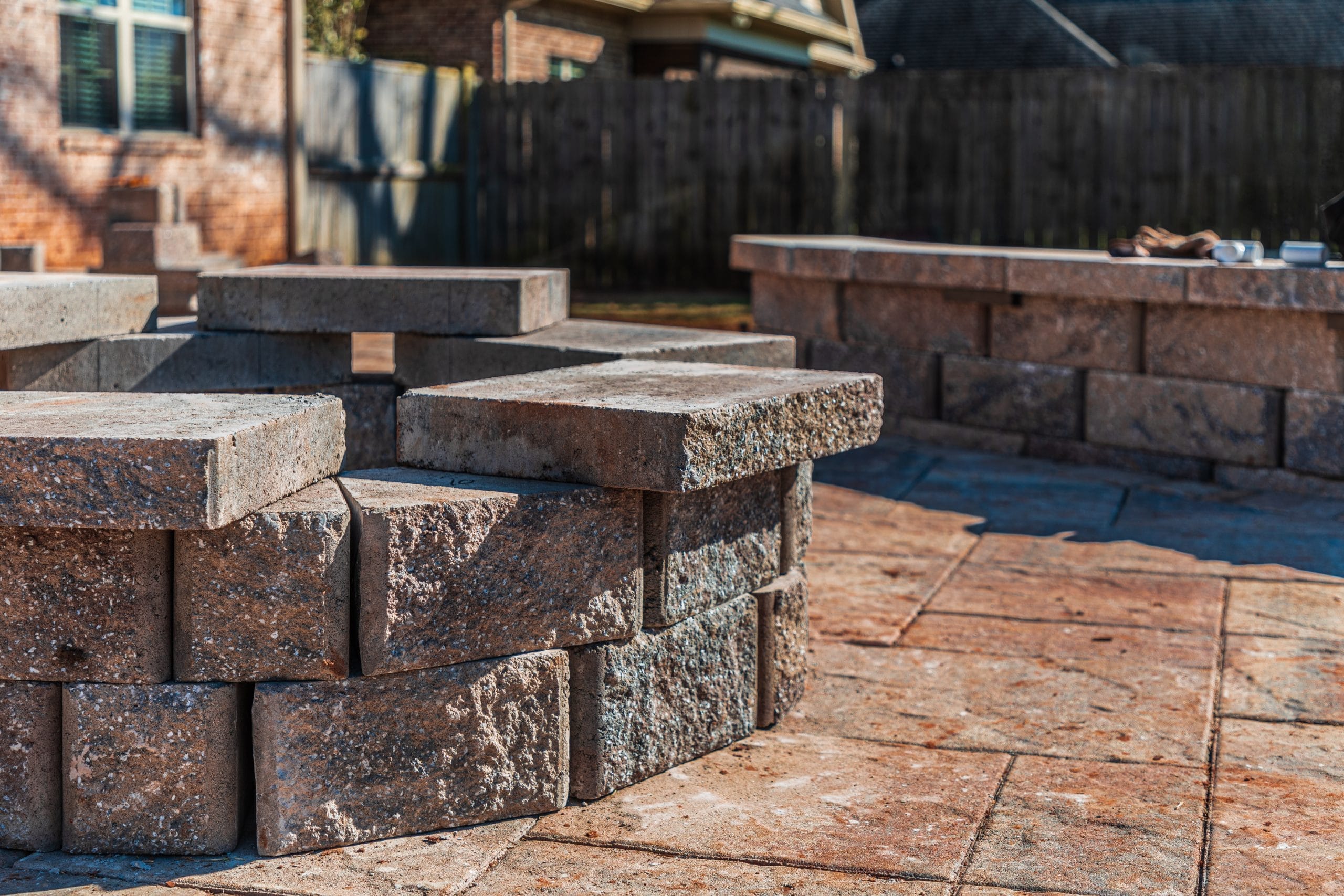 Close-up of a circular stone fire pit on a patio in a backyard with a brick house and wooden fence.