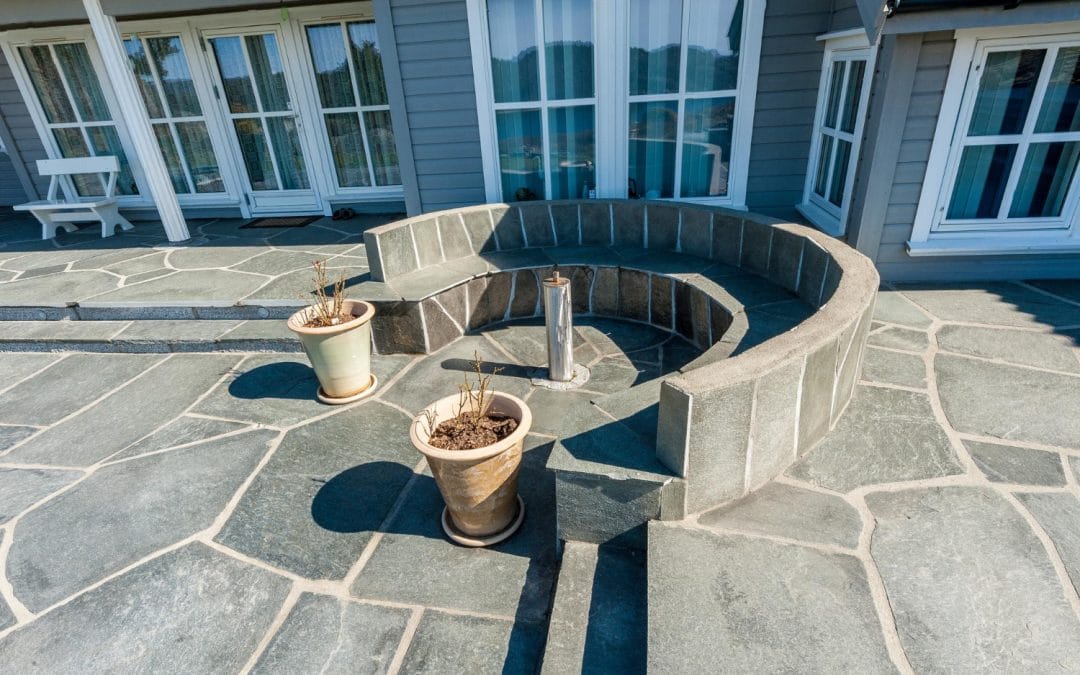 Curved stone bench and two potted plants on a spacious stone patio outside a house with large windows.
