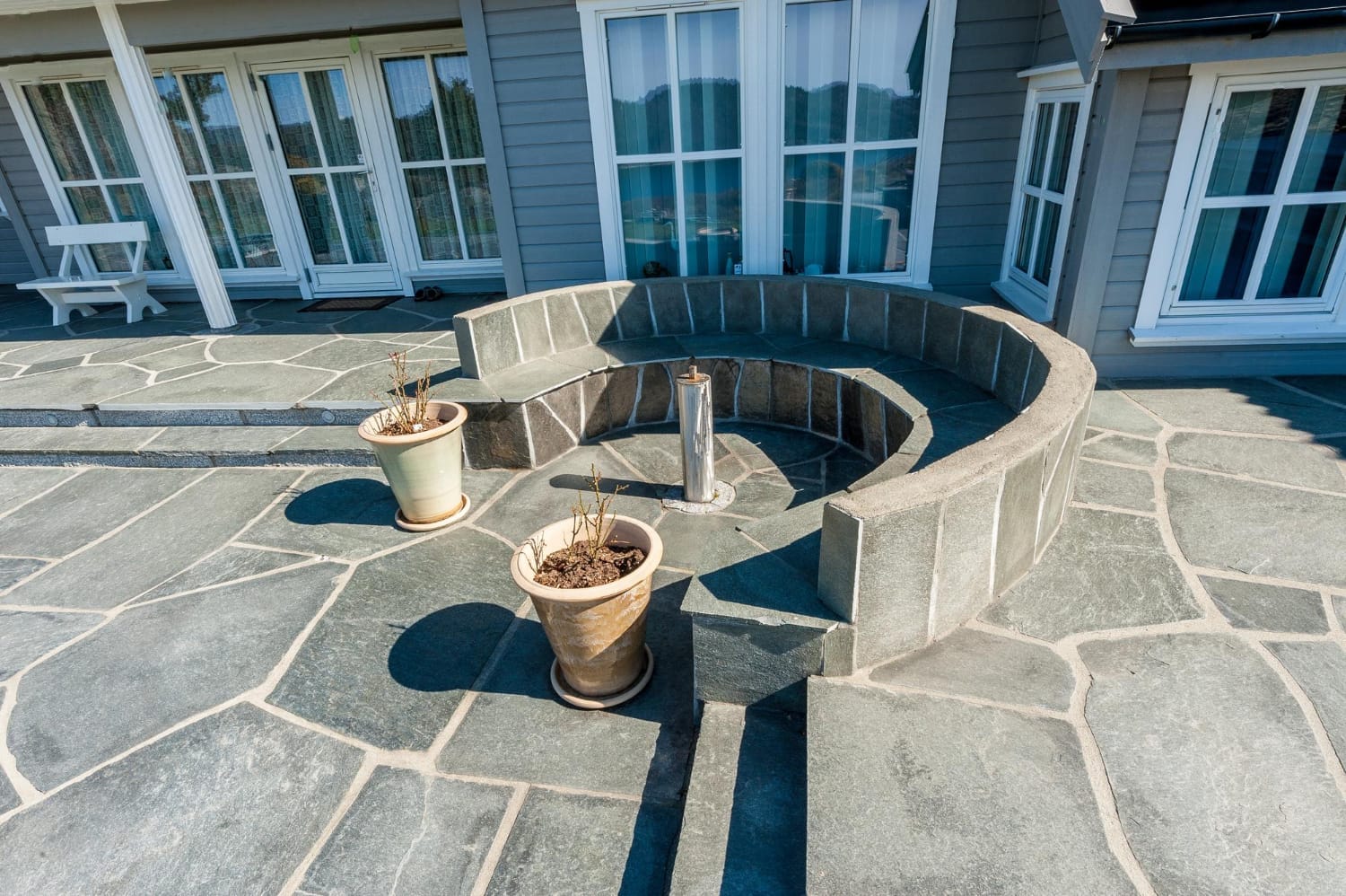 27156640 Curved stone bench and two potted plants on a spacious stone patio outside a house with large windows.