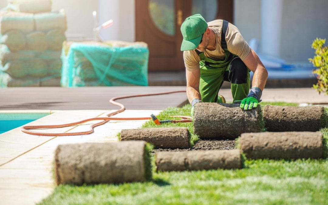 Worker in green uniform laying rolls of sod grass on a lawn next to a concrete patio.