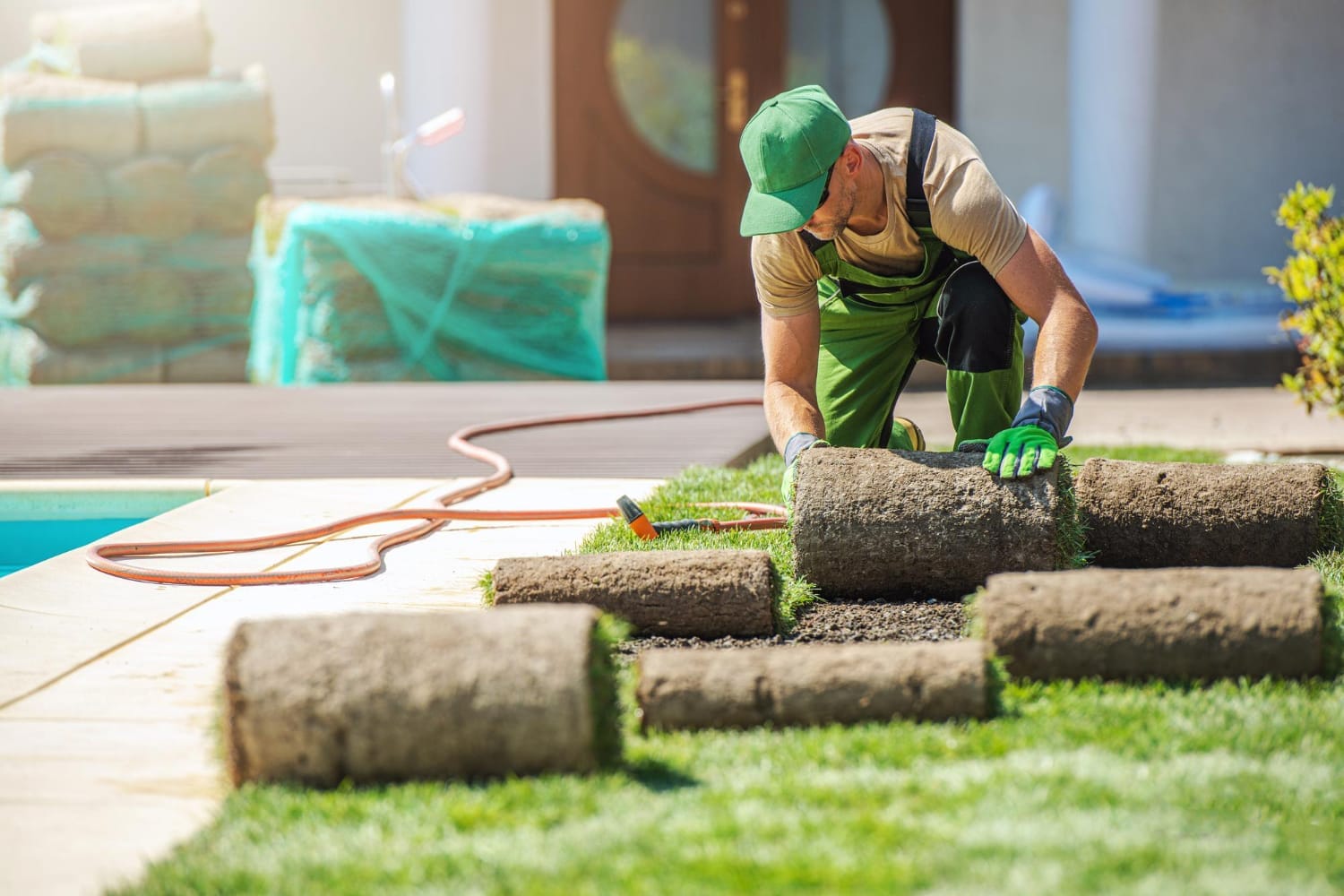 Worker in green uniform laying rolls of sod grass on a lawn next to a concrete patio.