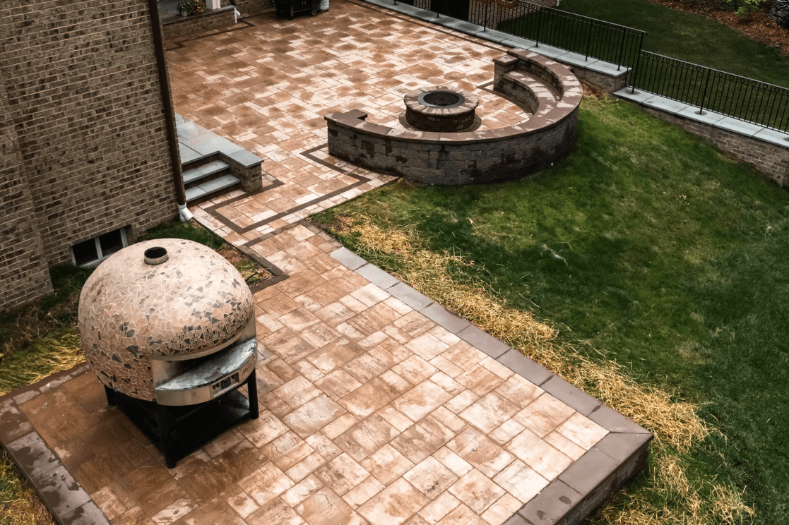 A backyard patio with a stone oven, fire pit, and curved stone seating, bordered by a grassy lawn.